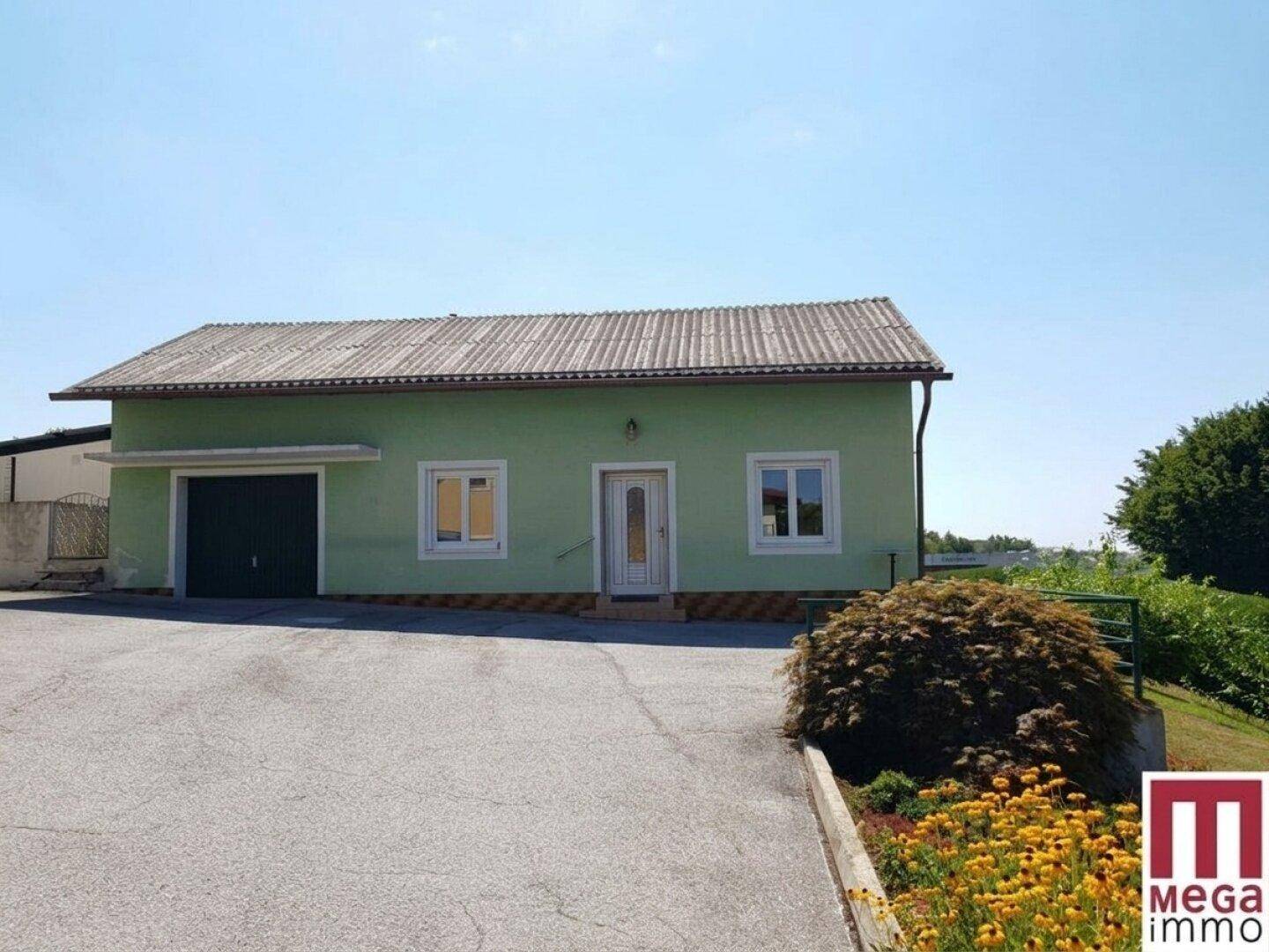 Einfamilienhaus mit grüner Fassade, integrierter Garage und gepflasterter Einfahrt unter blauem Himmel.