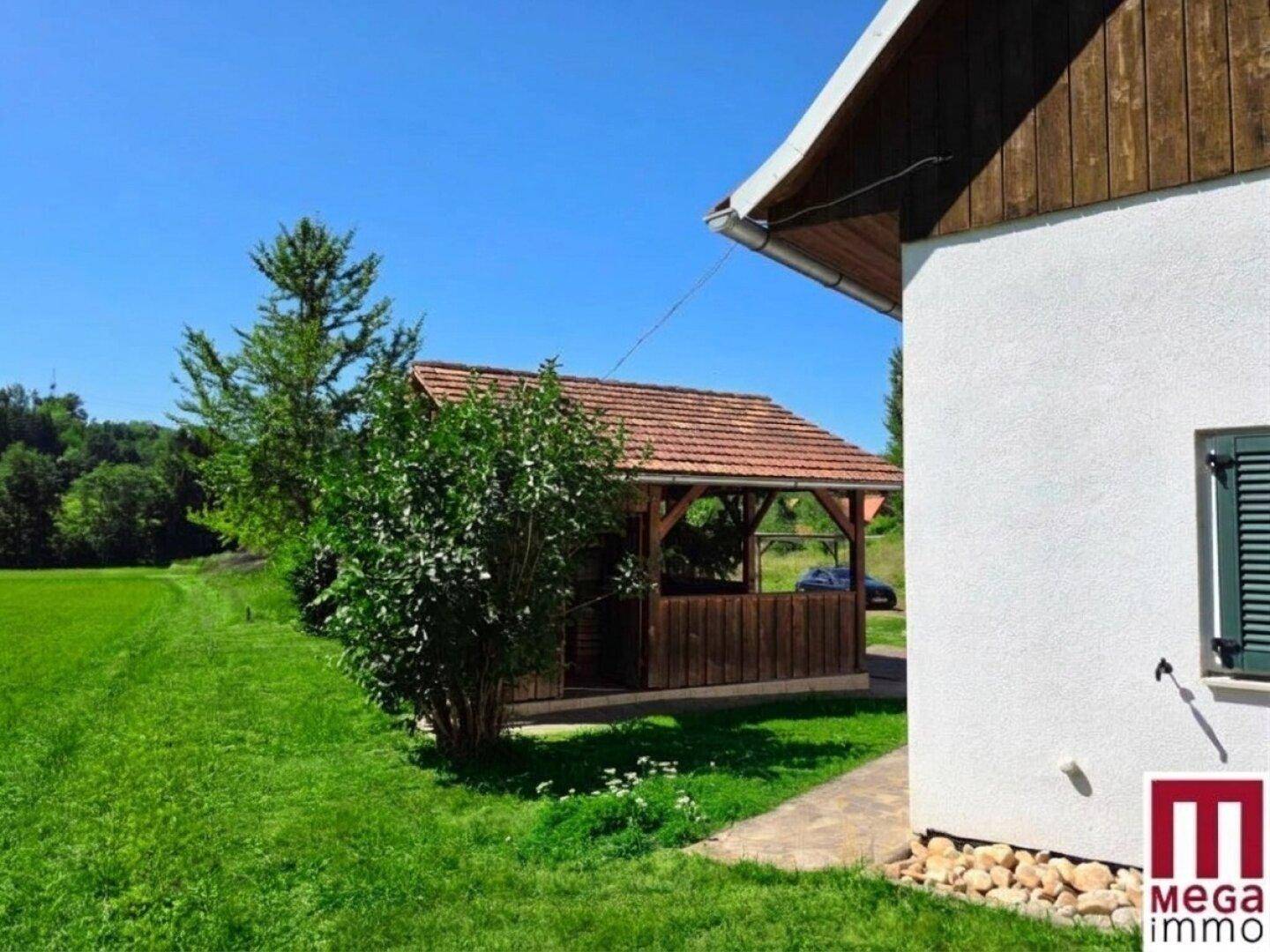 Seitenansicht des Hauses und des Holzpavillons, umgeben von grünen Feldern und einem gepflegten Garten.