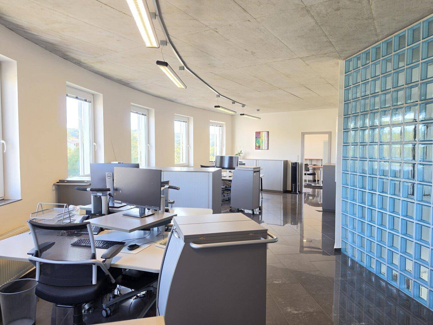 Offener Bürobereich mit Glasbausteinwand, mehreren Arbeitsplätzen und Fenstern mit Ausblick.