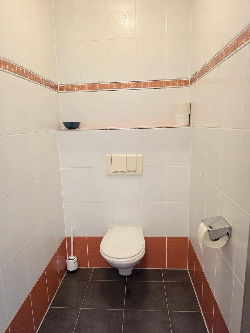 Gästetoilette mit wandhängendem WC und weiß-orange gefliesten Wänden und dunklem Boden.