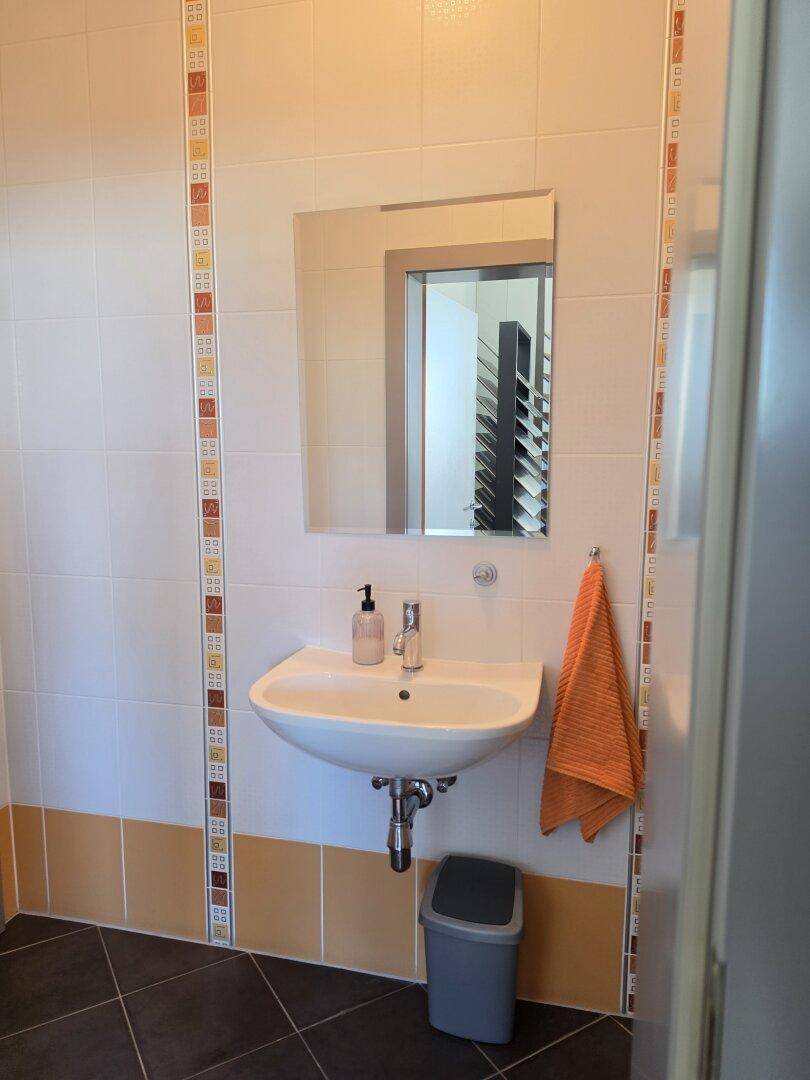 Gästetoilette mit Waschbecken, Spiegel und weiß-orange gefliesten Wänden und dunklem Boden.