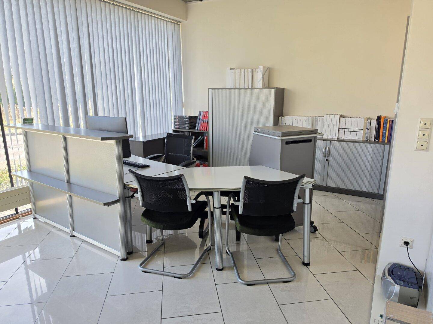Büroraum mit Schreibtisch, Bürostühlen und Aktenschränken, beleuchtet durch große Fenster mit Jalousien.