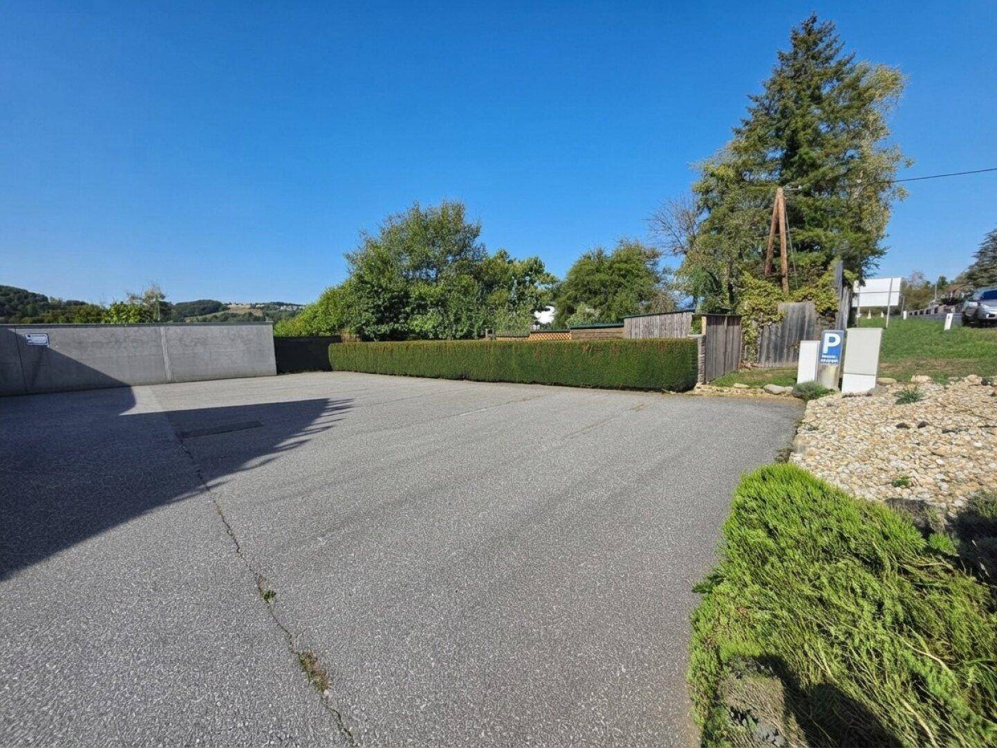 Großer asphaltierter Parkplatz mit Hecke und Bäumen im Hintergrund unter blauem Himmel.