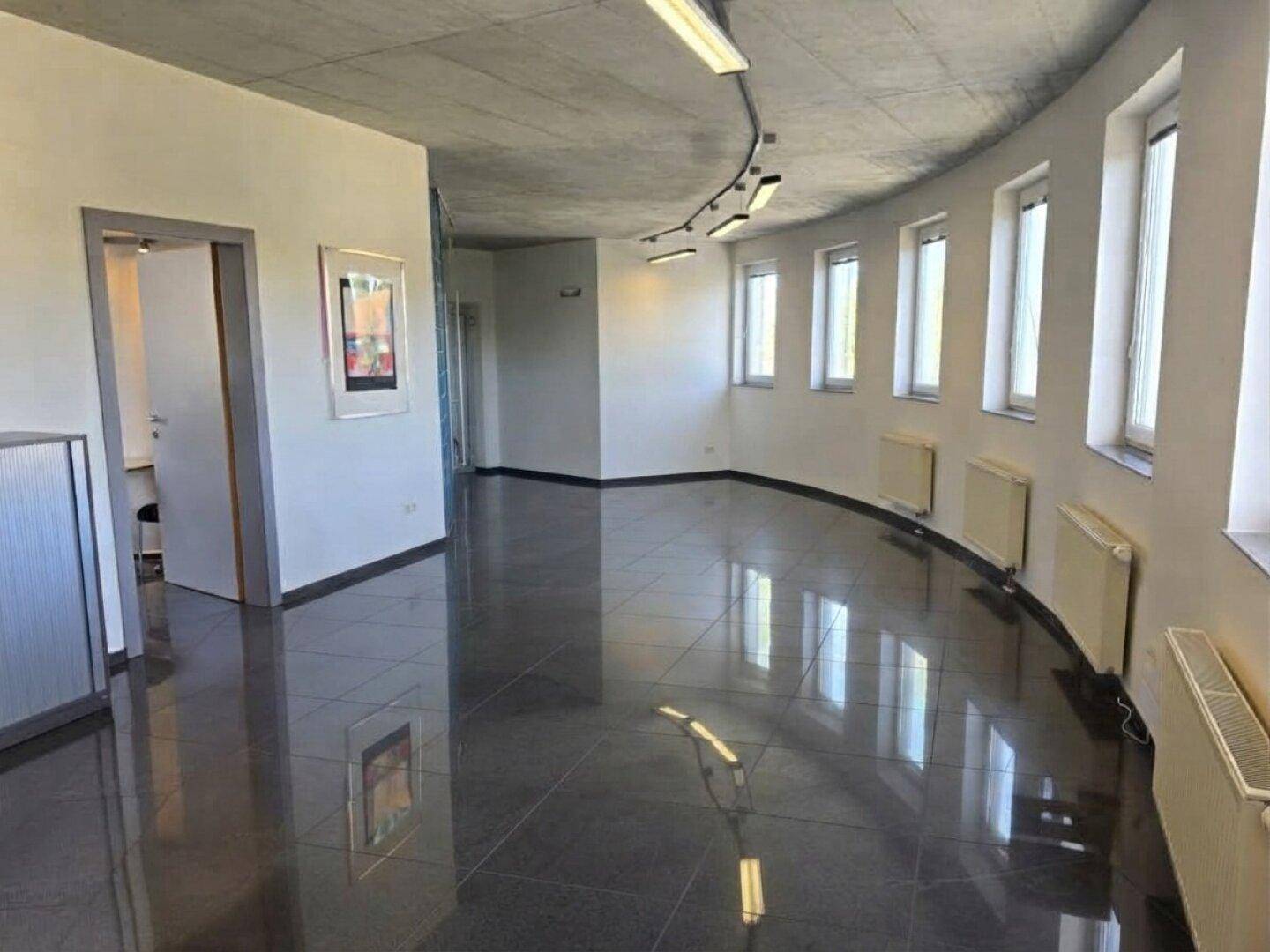 Leerer Bürobereich mit glänzenden dunklen Fliesen, Betonoptikdecke und Fenstern entlang der runden Wand.