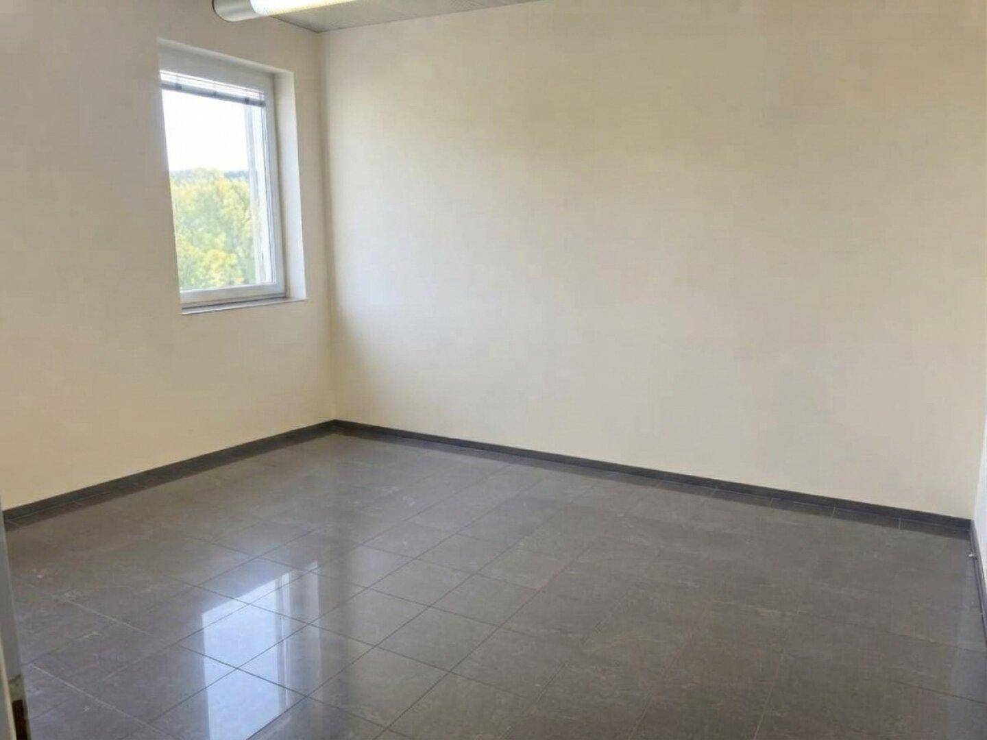 Leerer Büroraum mit hellen Wänden, dunklem Fliesenboden und einem Fenster, das Tageslicht spendet.