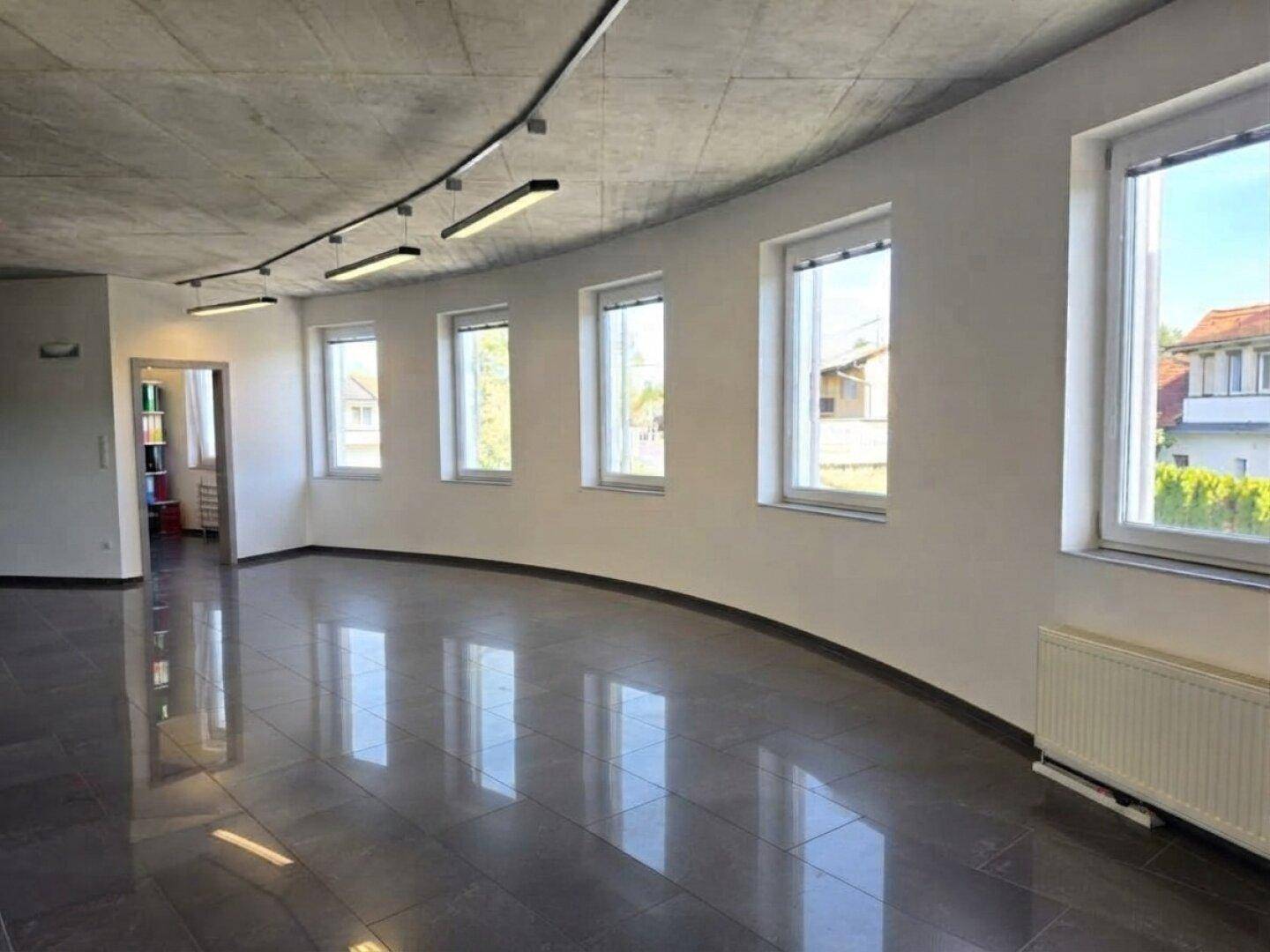 Geräumiger, leerer Bürobereich mit dunklen Fliesen, Betonoptikdecke und Fenstern entlang der runden Wand.