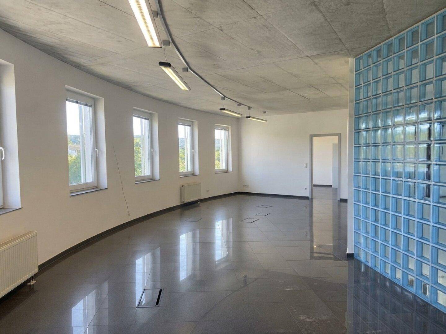 Großer, leerer Bürobereich mit Glasbausteinwand, dunklen Fliesen und Fenstern entlang der runden Wand.