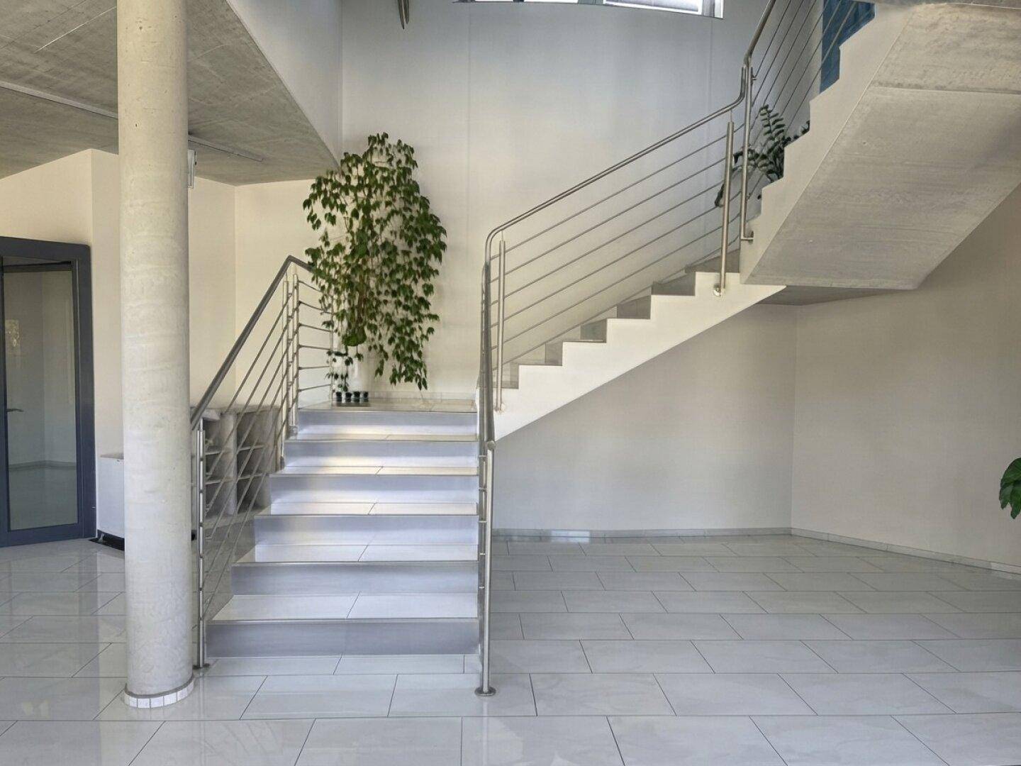 Heller Eingangsbereich mit moderner Treppe, Säule und großen Fenstern, die viel Licht hereinlassen.