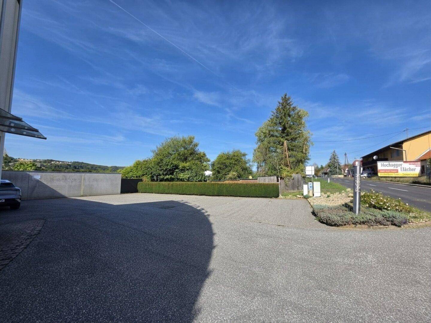Großer asphaltierter Parkplatz neben einer Straße mit Hecke und Bäumen im Hintergrund.