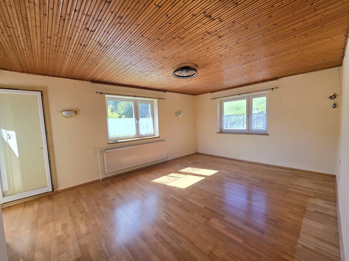 Geräumiges Wohnzimmer mit Holzboden, Holzdecke und zwei Fenstern.