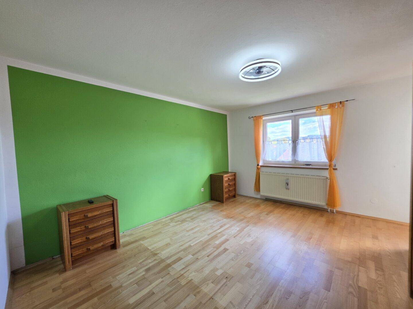 Schlafzimmer mit Holzboden, grüner Akzentwand und Fenster mit Vorhängen.