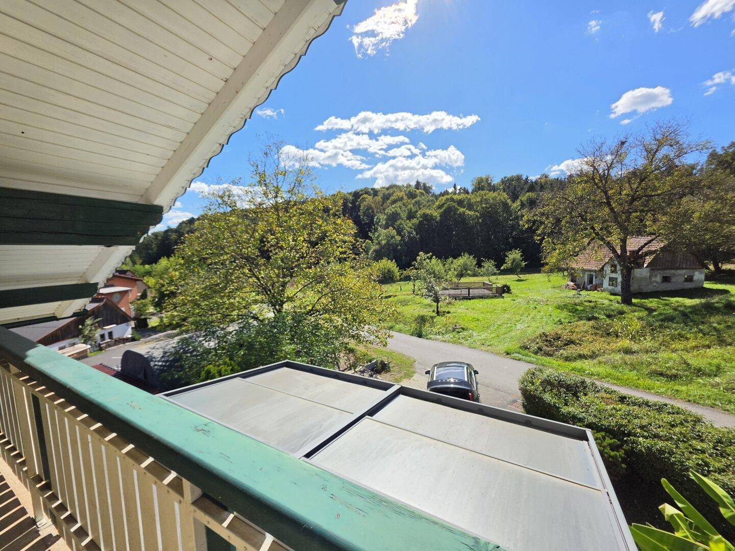 Weitläufiger Blick vom Balkon über grüne Felder, Bäume und ein kleines Landhaus unter blauem Himmel.