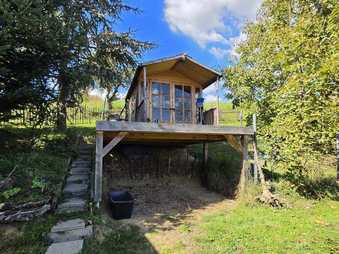 Rustikales Holzhaus auf Stelzen im Garten, umgeben von Bäumen und Grünflächen.