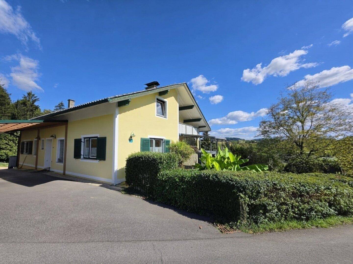 Seitenansicht des gelben Hauses mit Zufahrt, Hecke und ländlicher Umgebung.