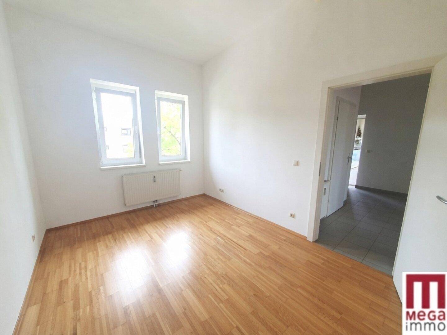 Leeres Zimmer mit Parkettboden, weißen Wänden und zwei Fenstern für viel Tageslicht.