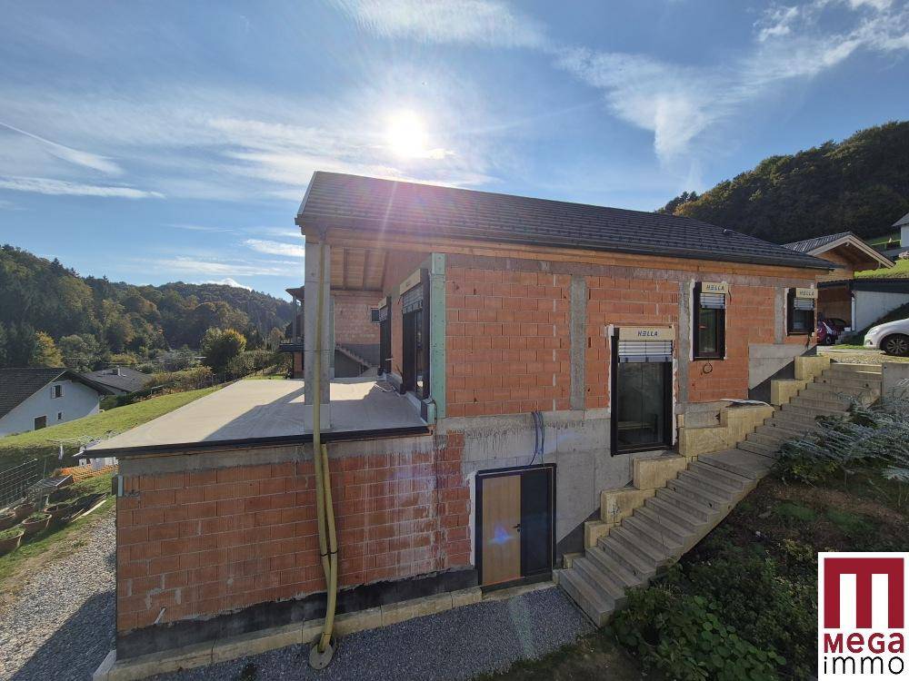 Haus im Bau mit Ziegelwänden, Fensterrahmen und Rollläden, umgeben von Natur.