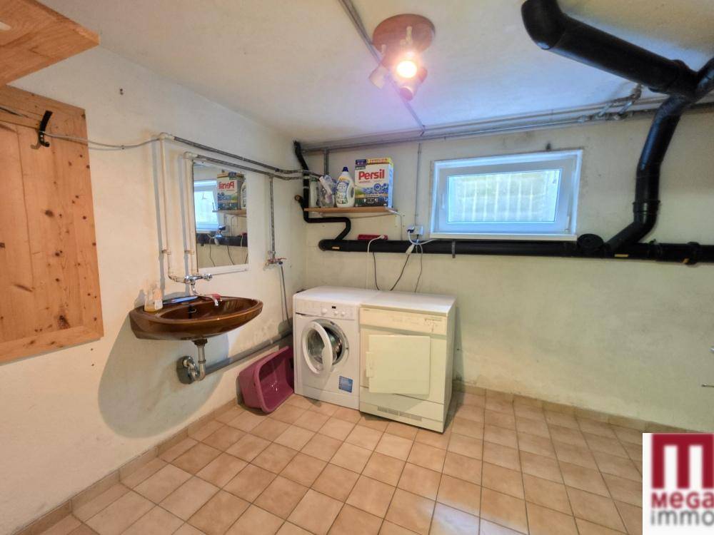 Waschküche im Keller mit Waschmaschine, Trockner, Waschbecken und sichtbaren Rohren an der Wand.