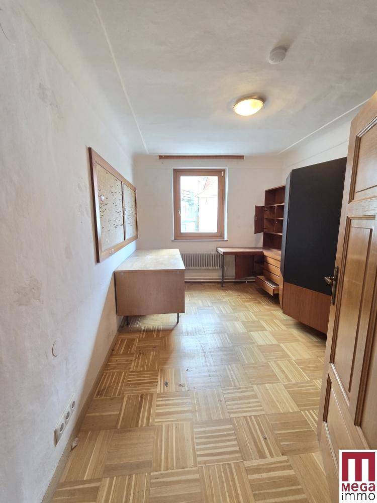 Helles Zimmer mit Parkettboden, Fenster und einfacher Einrichtung.