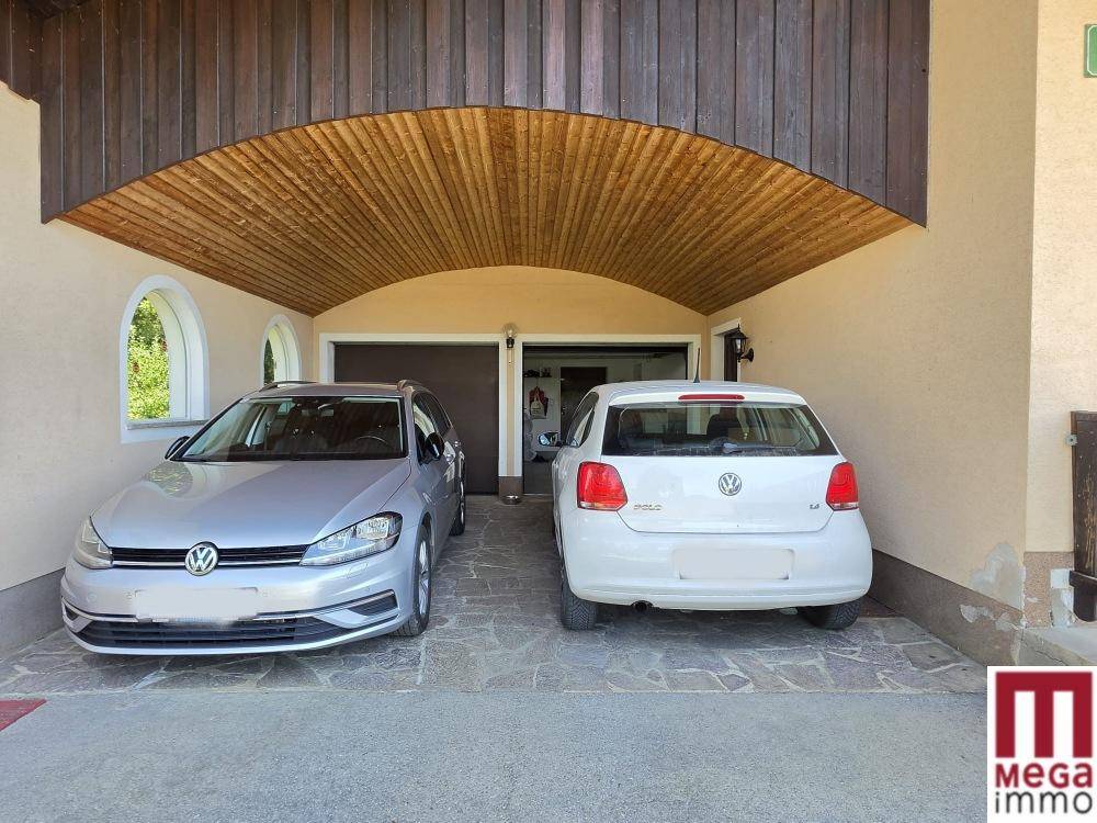Überdachter Stellplatz für zwei Fahrzeuge vor der Garage mit Holzbalkendecke und gepflastertem Boden.
