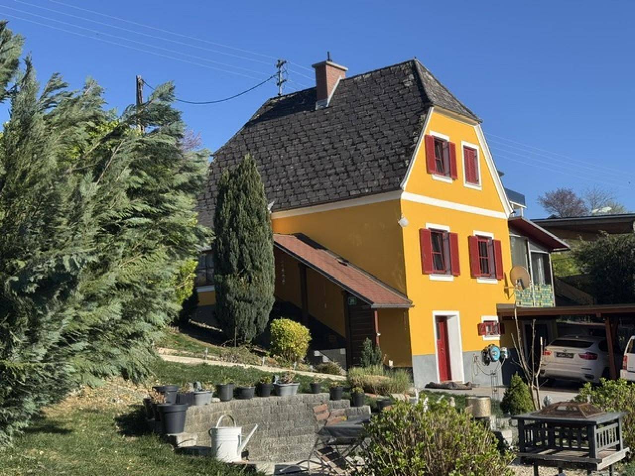 Nahaufnahme des gelben Hauses mit traditionellem Dach und roten Fensterläden, umgeben von Grünflächen.