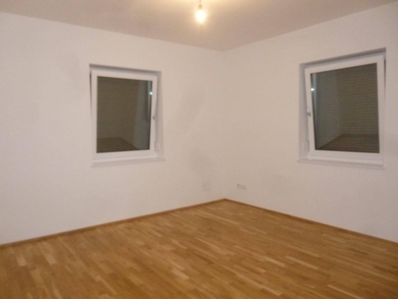 Helles leeres Zimmer mit Holzparkettboden und zwei Fenstern.