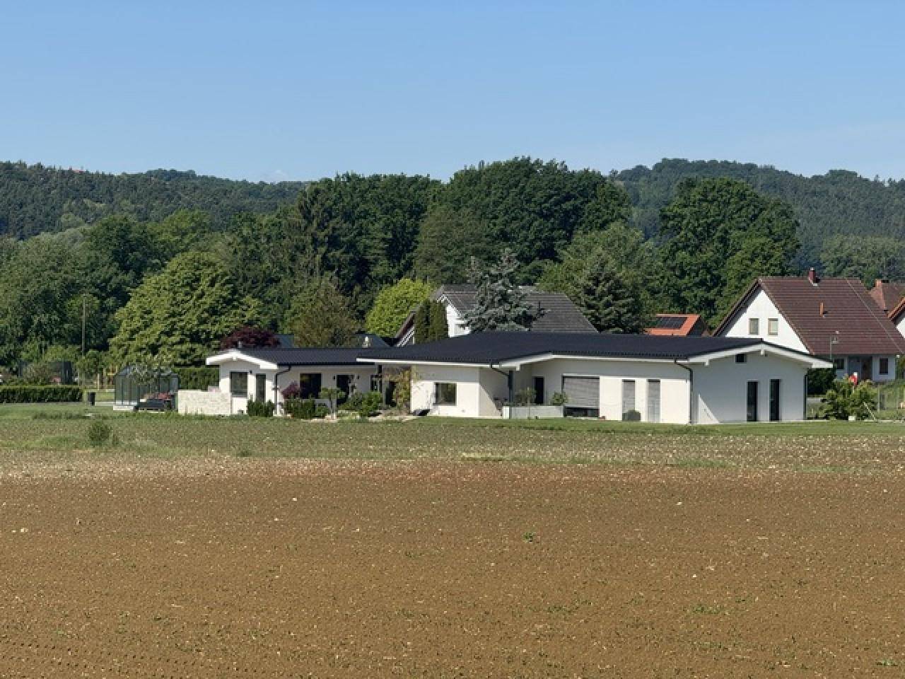 Modernes Einfamilienhaus mit weißer Fassade und dunklem Dach, eingebettet in eine grüne Landschaft.