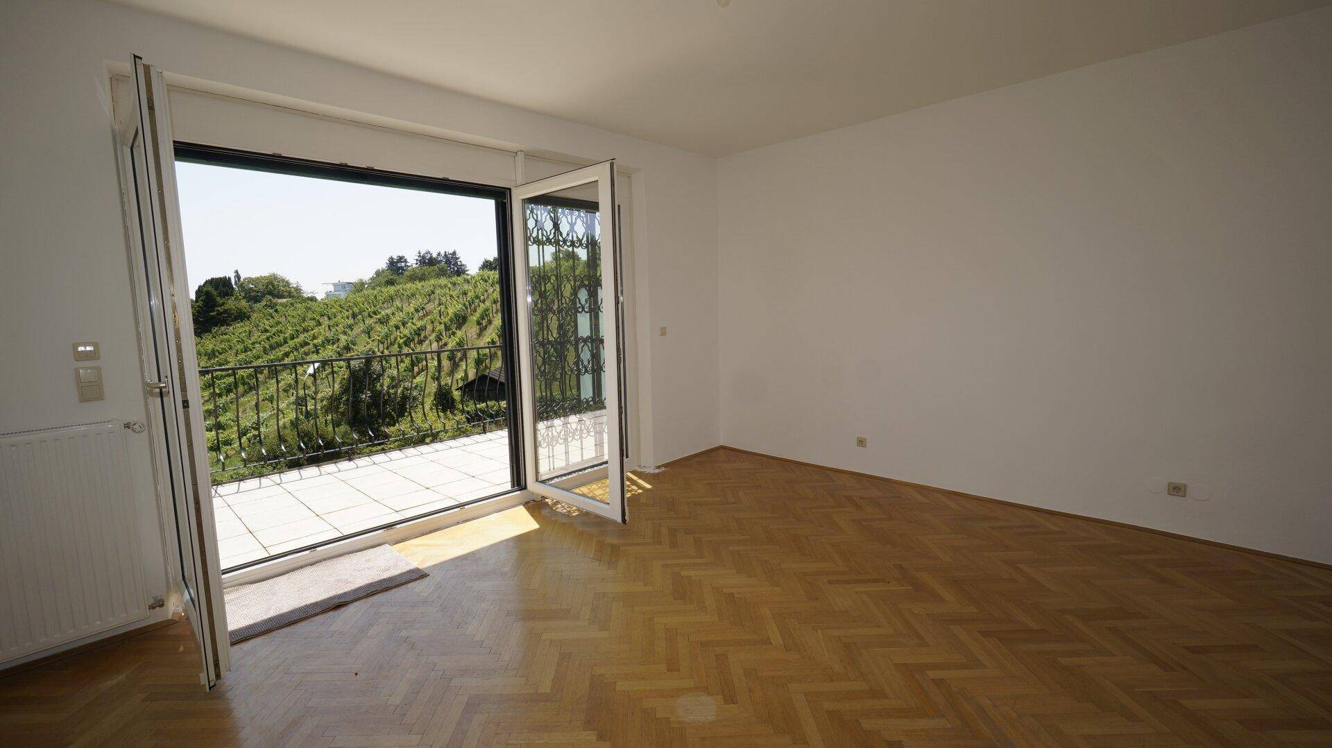 Heller Raum mit Fischgrätparkett und Zugang zu einem Balkon mit Weinbergblick.