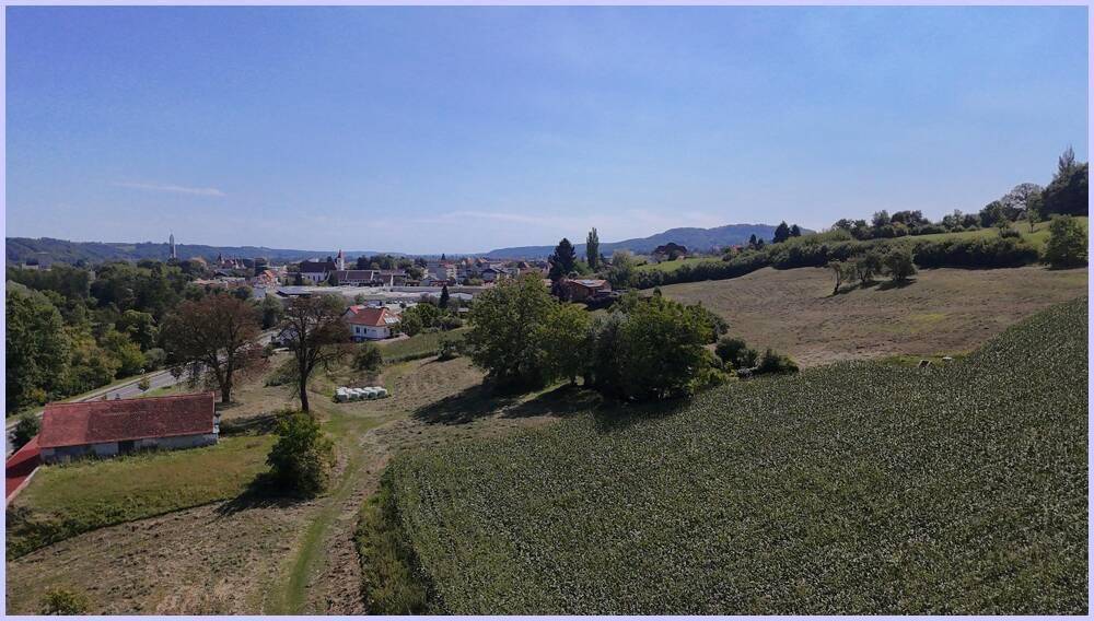 Weitläufige Landschaftsaufnahme mit einem Feld im Vordergrund und einem Dorf am Horizont unter blauem Himmel.