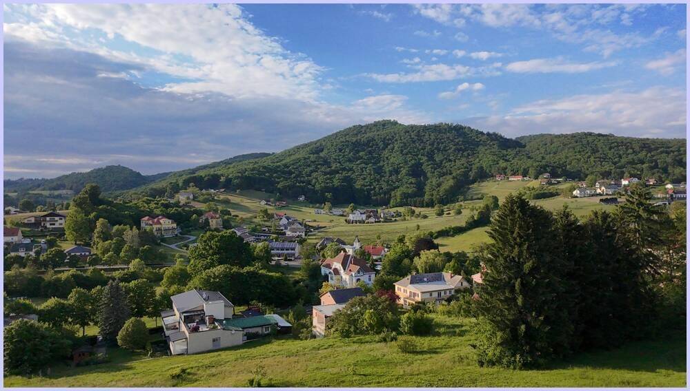 Weitläufige Landschaft mit grünen Hügeln, Wäldern und verstreuten Häusern unter blauem Himmel.
