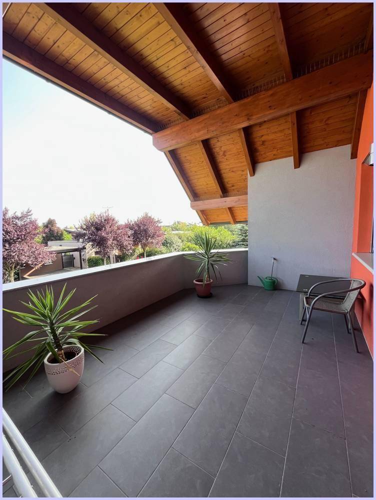 Großer überdachter Balkon mit Holzdach, Fliesenboden und Blick ins Grüne.