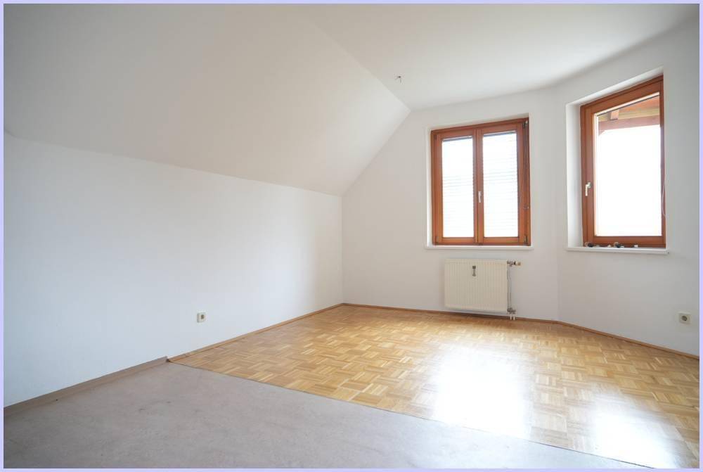 Heller Raum mit Parkettboden, Heizkörper und zwei Fenstern unter einer Dachschräge.