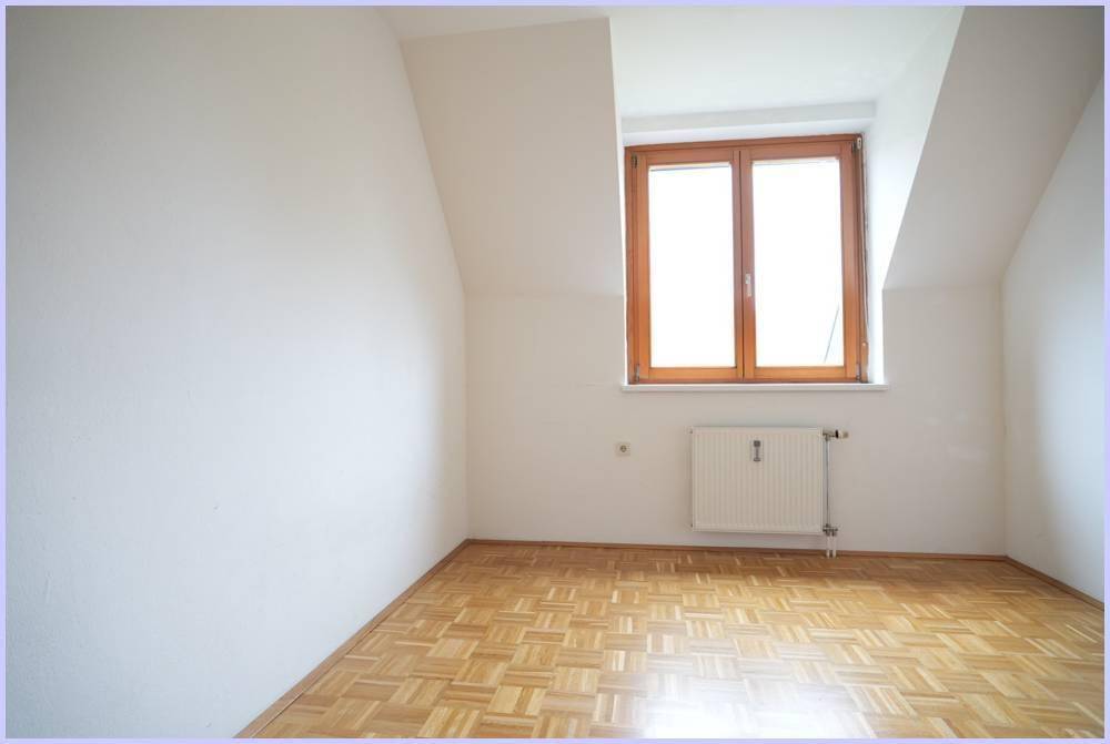 Heller Raum mit Parkettboden, Heizkörper und Fenster unter einer Dachschräge.