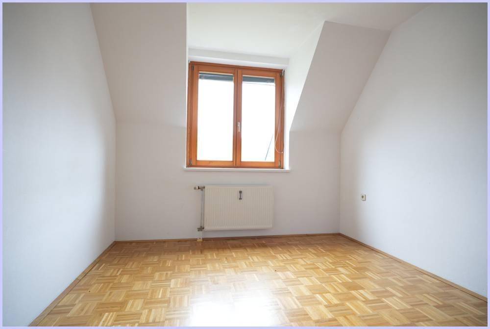 Leerer Raum mit Parkettboden, Heizkörper und Fenster unter einer Dachschräge.