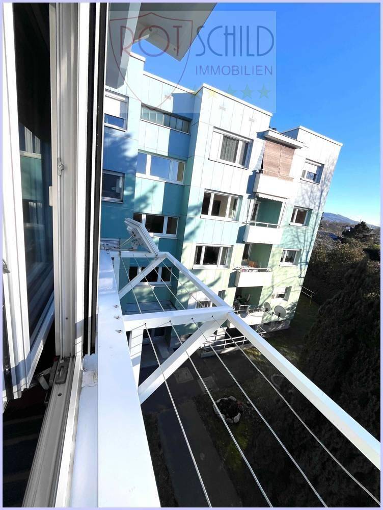 Blick vom Balkon auf eine Wäscheleine und ein Mehrfamilienhaus mit blauer Fassade und Bergblick.