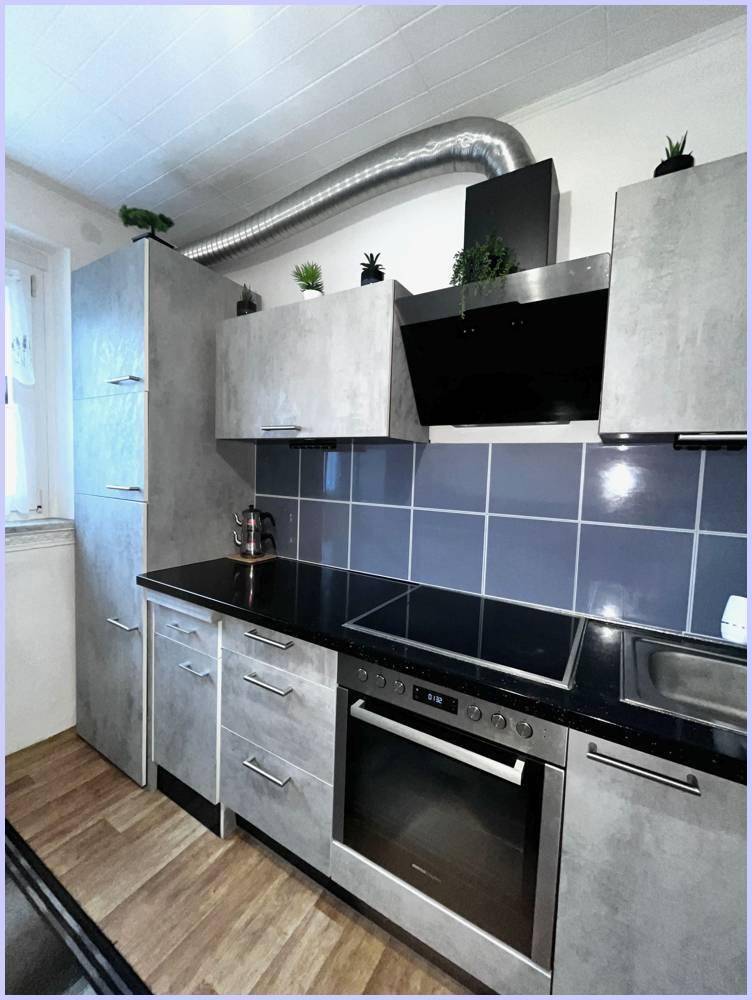 Moderne Küche mit grauen Schränken, schwarzer Arbeitsplatte und integriertem Backofen und Kochfeld.