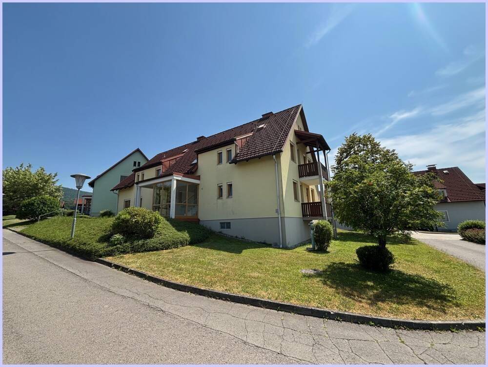 Mehrfamilienhaus mit Balkonen und Wintergarten in grüner Umgebung unter blauem Himmel.