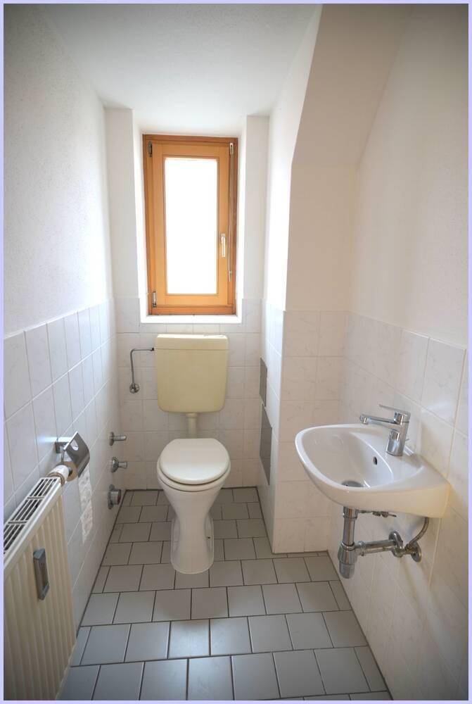 Separates WC mit Toilette, kleinem Waschbecken und Fenster, hell gefliest.