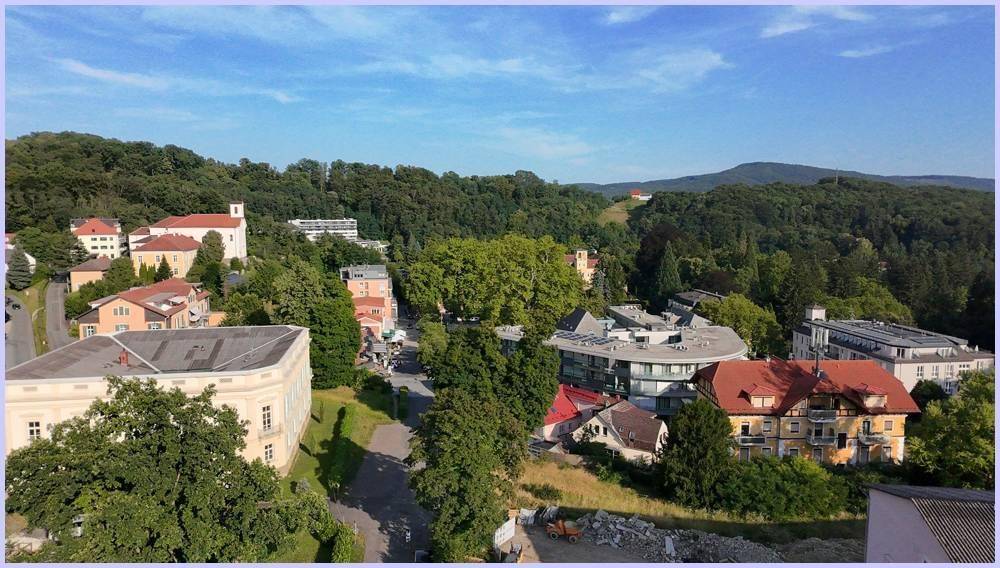 Panoramablick über eine grüne Hügellandschaft mit Wohngebäuden und Wald unter blauem Himmel.