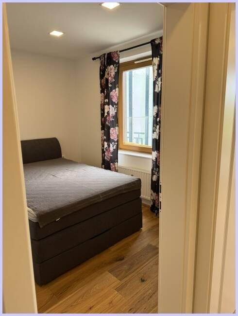 Schlafzimmer mit dunklem Bett, floralen Vorhängen und Fenster, das Tageslicht hereinlässt.