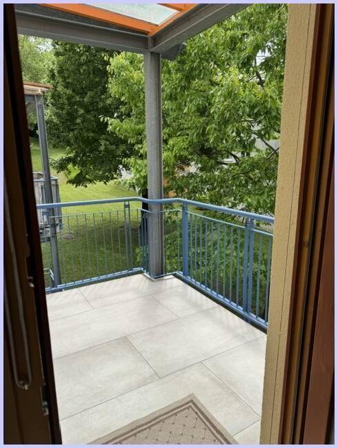 Überdachter Balkon mit Fliesenboden und blauem Geländer, umgeben von grünen Bäumen.