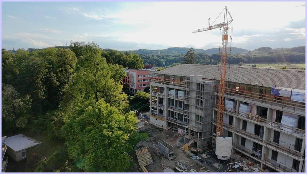 Baustelle eines Neubaus mit Kran, umgeben von dichtem Grün und weiteren Gebäuden im Hintergrund.