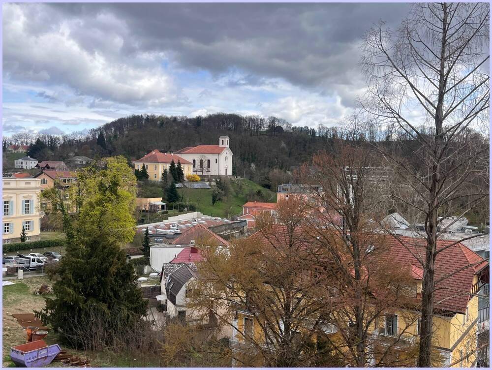 Blick auf eine Stadt mit einer Kirche auf einem Hügel, umgeben von Bäumen unter bewölktem Himmel.