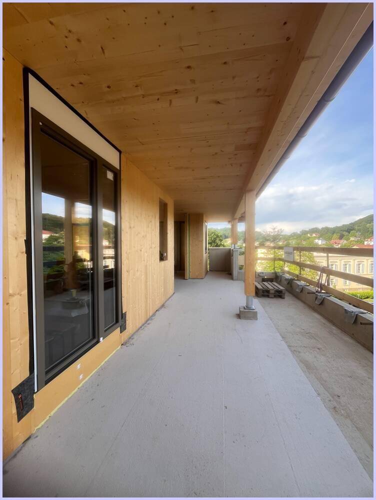 Langer Balkon im Bau mit Holzdecke und großen Fenstern, mit Blick auf entfernte Gebäude.