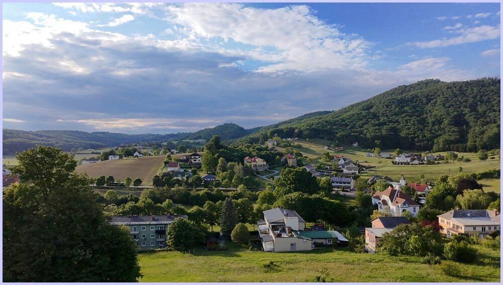 Panoramablick auf eine grüne, hügelige Landschaft mit verstreuten Häusern unter teilweise bewölktem Himmel.