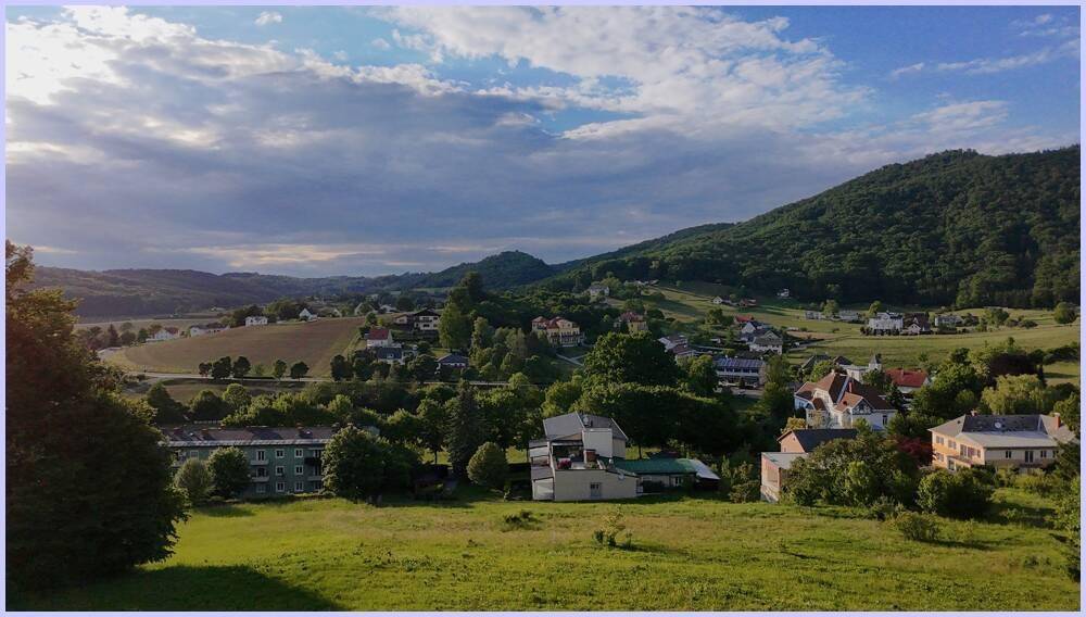 Panoramablick auf eine grüne, hügelige Landschaft mit verstreuten Häusern unter bewölktem Himmel.
