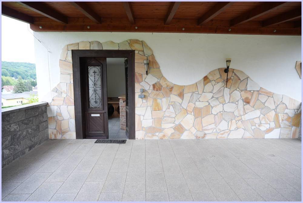 Geräumige überdachte Terrasse mit Natursteinwand und Zugang zum Haus, ideal für Entspannung.