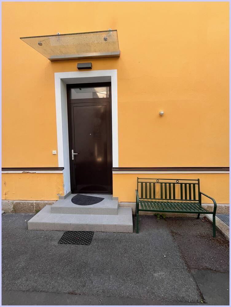 Detaillierter Blick auf den Hauseingang mit dunkler Tür, Glasvordach und Sitzbank.
