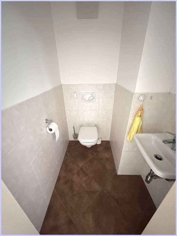 Separates WC mit wandhängender Toilette, kleinem Waschbecken und Fliesenboden.