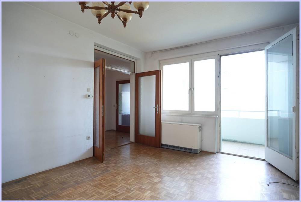 Geräumiges Wohnzimmer mit Parkettboden, Zugang zum Balkon und einem Heizkörper unter dem Fenster.
