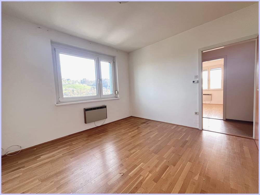 Helles Zimmer mit Holzboden und Blick ins Grüne, verbunden mit einem weiteren Raum.